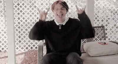 A Young Man In A Black Sweater Is Sitting In A Chair And Making A Devil Horns Sign . GIF