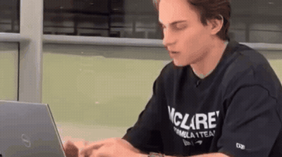 A Young Man In A Black T-Shirt Is Sitting At A Table Using A Laptop Computer . GIF