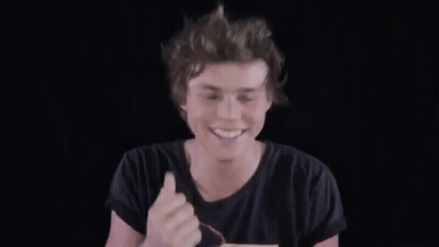 A Young Man In A Black T-Shirt Is Smiling And Waving His Hand In The Air . GIF