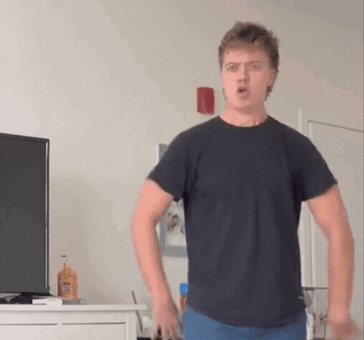 A Young Man In A Black T-Shirt Is Standing In Front Of A Television In A Living Room . GIF