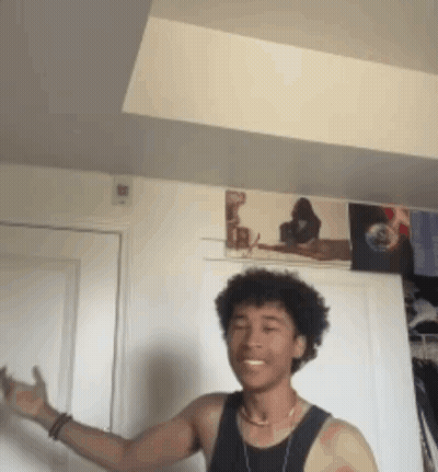 A Young Man In A Black Tank Top Is Standing In A Room With A Picture On The Wall Behind Him . GIF
