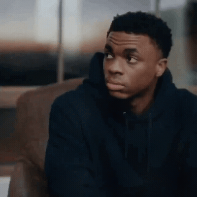 A Young Man In A Blue Hoodie Is Sitting On A Couch And Looking At The Camera . GIF