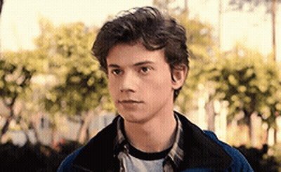 A Young Man In A Blue Jacket And Plaid Shirt Is Standing In Front Of Trees . GIF