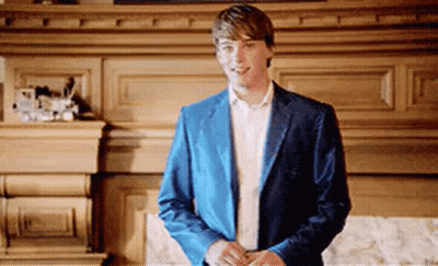 A Young Man In A Blue Jacket And White Shirt Is Standing In Front Of A Fireplace . GIF