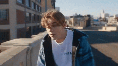 A Young Man In A Blue Jacket Is Standing On A Bridge . GIF