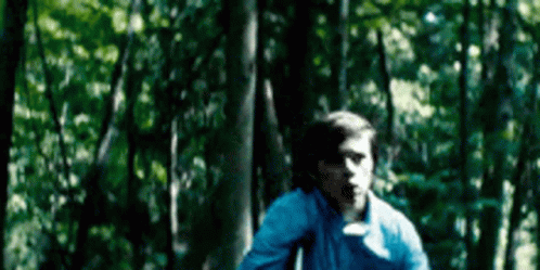 A Young Man In A Blue Shirt Is Running Through A Forest . GIF