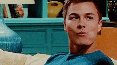A Young Man In A Blue Shirt Is Sitting On A Couch And Making A Funny Face . GIF