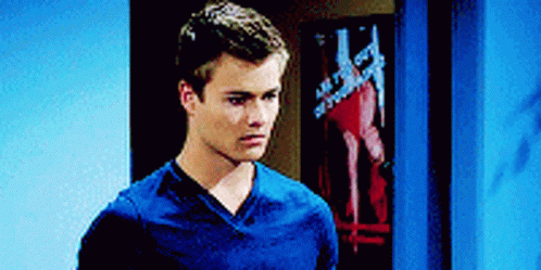 A Young Man In A Blue Shirt Is Standing In Front Of A Door GIF