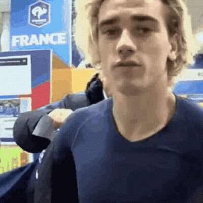 A Young Man In A Blue Shirt Is Standing In Front Of A France Sign . GIF