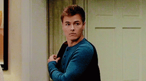 A Young Man In A Blue Sweater Is Standing In Front Of A Door Holding His Jacket . GIF