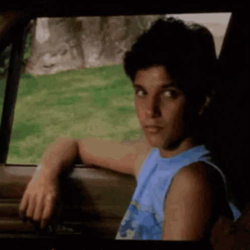 A Young Man In A Blue Tank Top Is Leaning On The Door Of A Car . GIF