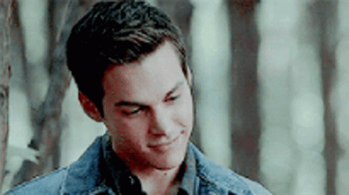 A Young Man In A Denim Jacket Is Smiling In Front Of A Forest . GIF