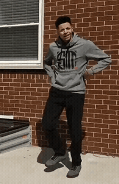 A Young Man In A Gray Hoodie Is Standing In Front Of A Brick Building . GIF