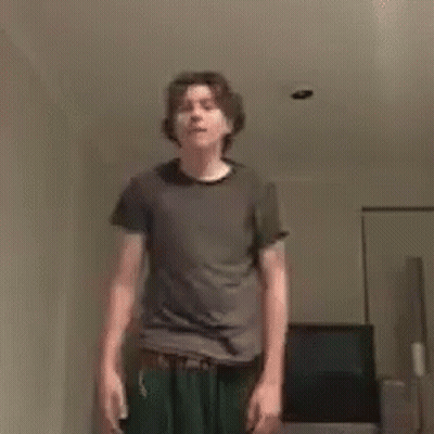 A Young Man In A Gray Shirt And Green Pants Is Standing In A Living Room . GIF