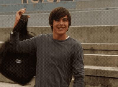 A Young Man In A Gray Shirt Is Holding A Black Bag With The Letter T On It GIF