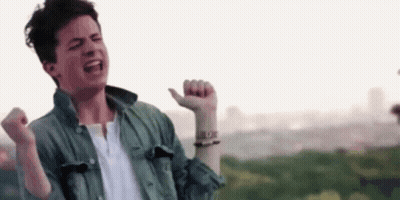 A Young Man In A Green Jacket And White Shirt Is Standing In Front Of A City With His Arms In The Air . GIF