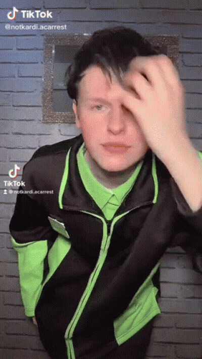 A Young Man In A Green Shirt And Black Jacket Is Standing In Front Of A Brick Wall . GIF