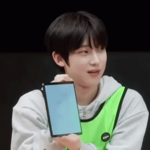 A Young Man In A Green Vest Is Holding A Cell Phone . GIF