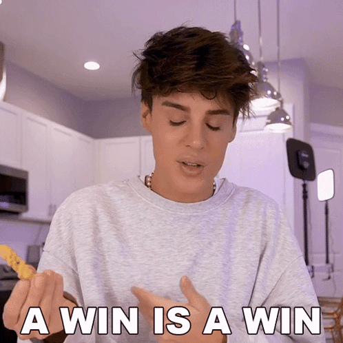 A Young Man In A Grey Sweater Says " A Win Is A Win " In A Kitchen GIF