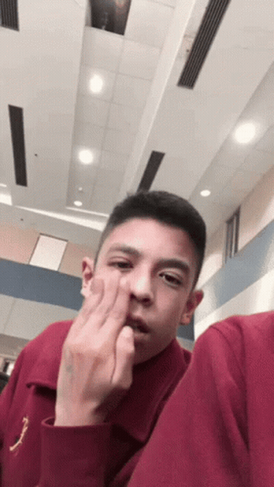 A Young Man In A Maroon Shirt Is Making A Funny Face While Sitting In A Room . GIF