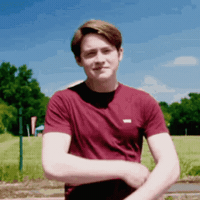 A Young Man In A Maroon Shirt Is Standing In A Field With His Arms Crossed . GIF