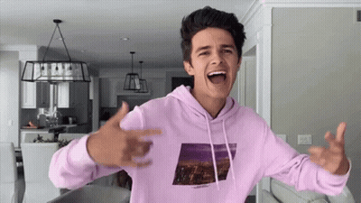 A Young Man In A Pink Hoodie Is Standing In A Living Room With His Arms Outstretched . GIF