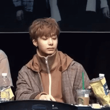 A Young Man In A Plaid Jacket Is Sitting At A Table Holding A Cup Of Coffee And A Bag Of Chips . GIF