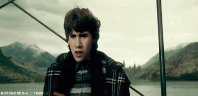 A Young Man In A Plaid Jacket Is Standing Next To A Body Of Water With Mountains In The Background . GIF