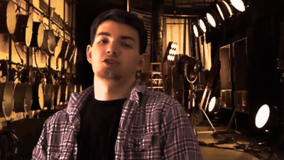 A Young Man In A Plaid Shirt Stands In Front Of A Wall Of Drums GIF