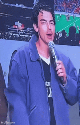 A Young Man In A Purple Jacket Is Holding A Microphone And Talking Into It . GIF
