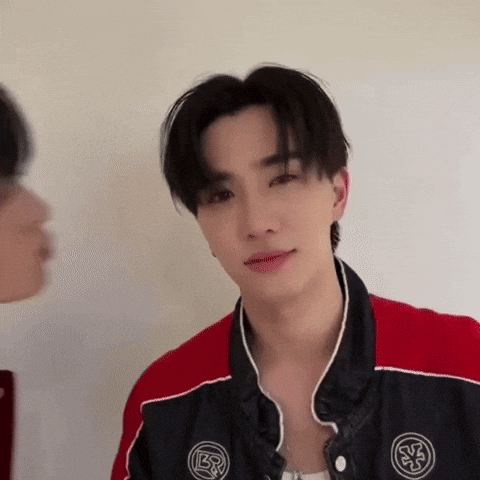 A Young Man In A Red And Black Jacket Is Looking At The Camera While Another Man Looks On . GIF