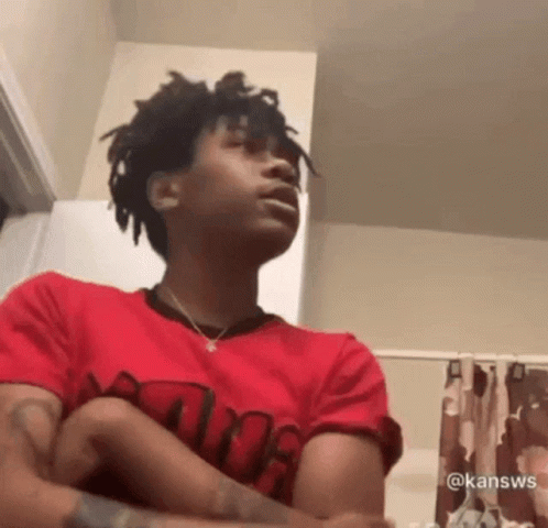 A Young Man In A Red Shirt Is Sitting In A Bathroom With His Arms Crossed And His Mouth Open . GIF