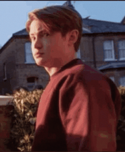 A Young Man In A Red Sweater Is Standing In Front Of A House And Looking At The Camera . GIF