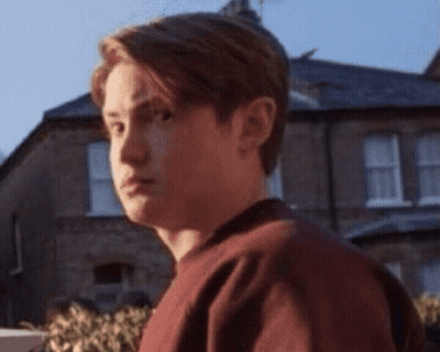 A Young Man In A Red Sweater Is Standing In Front Of A House . GIF