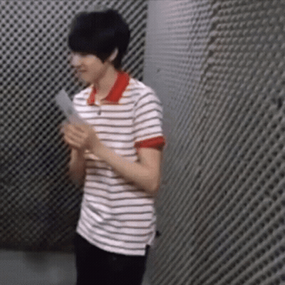 A Young Man In A Striped Shirt Is Standing In Front Of A Grid Wall Holding A Piece Of Paper . GIF