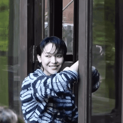 A Young Man In A Striped Sweater Is Smiling While Standing In A Doorway . GIF