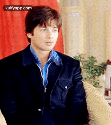A Young Man In A Suit And Blue Shirt Is Sitting In A Chair . GIF