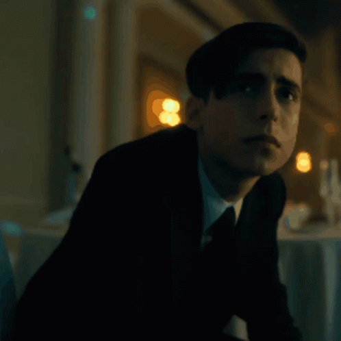 A Young Man In A Suit And Tie Is Sitting In A Dark Room . GIF