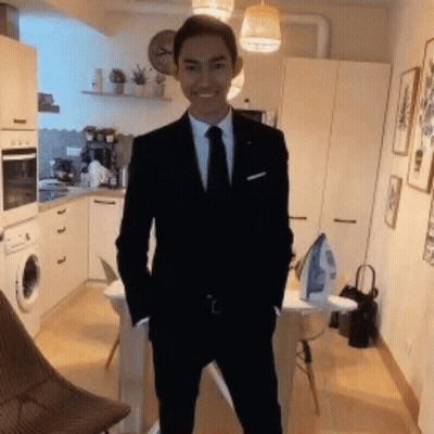 A Young Man In A Suit And Tie Is Standing In A Kitchen With His Hands In His Pockets . GIF