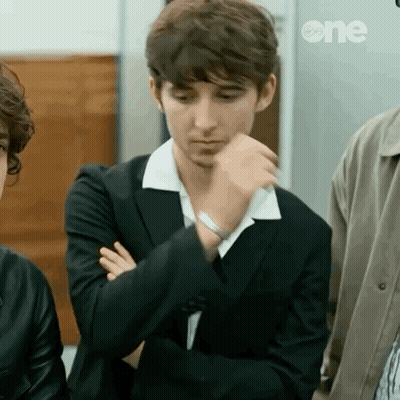 A Young Man In A Suit And White Collar Is Sitting With His Arms Crossed And His Hand On His Chin . GIF