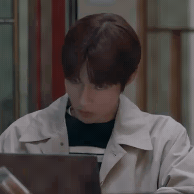A Young Man In A Trench Coat Is Sitting In Front Of A Laptop . GIF