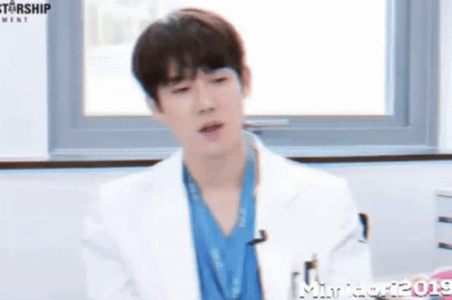 A Young Man In A White Coat And Blue Scrub Is Sitting In Front Of A Window . GIF