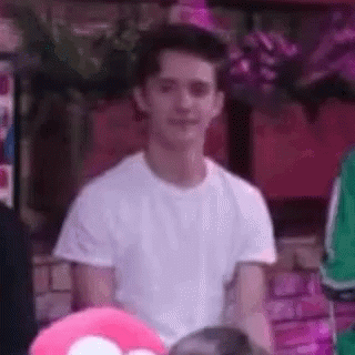A Young Man In A White T-Shirt Is Sitting In Front Of A Pink Heart Pillow . GIF