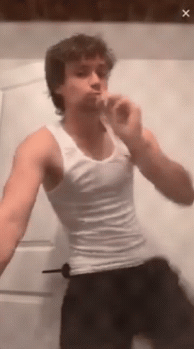 A Young Man In A White Tank Top Is Brushing His Teeth In A Bathroom . GIF