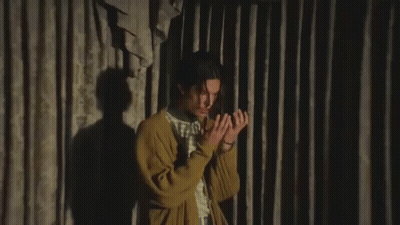 A Young Man In A Yellow Cardigan Is Making A Funny Face While Sitting In Front Of A Curtain . GIF