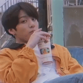 A Young Man In A Yellow Jacket Is Sitting On A Bench Drinking From A Cup . GIF