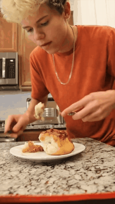 A Young Man In An Orange Shirt Is Eating A Cinnamon Roll GIF