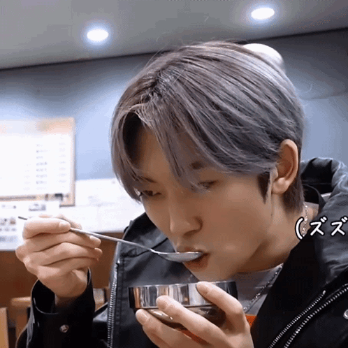 A Young Man Is Eating From A Bowl With A Spoon While Wearing A Black Jacket GIF