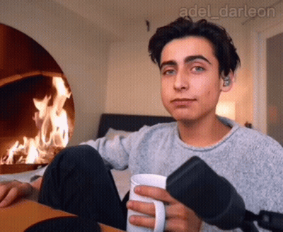 A Young Man Is Holding A Cup Of Coffee In Front Of A Fireplace GIF