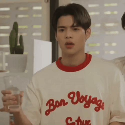 A Young Man Is Holding A Glass Of Water While Wearing A T-Shirt That Says Bon Voyage . GIF